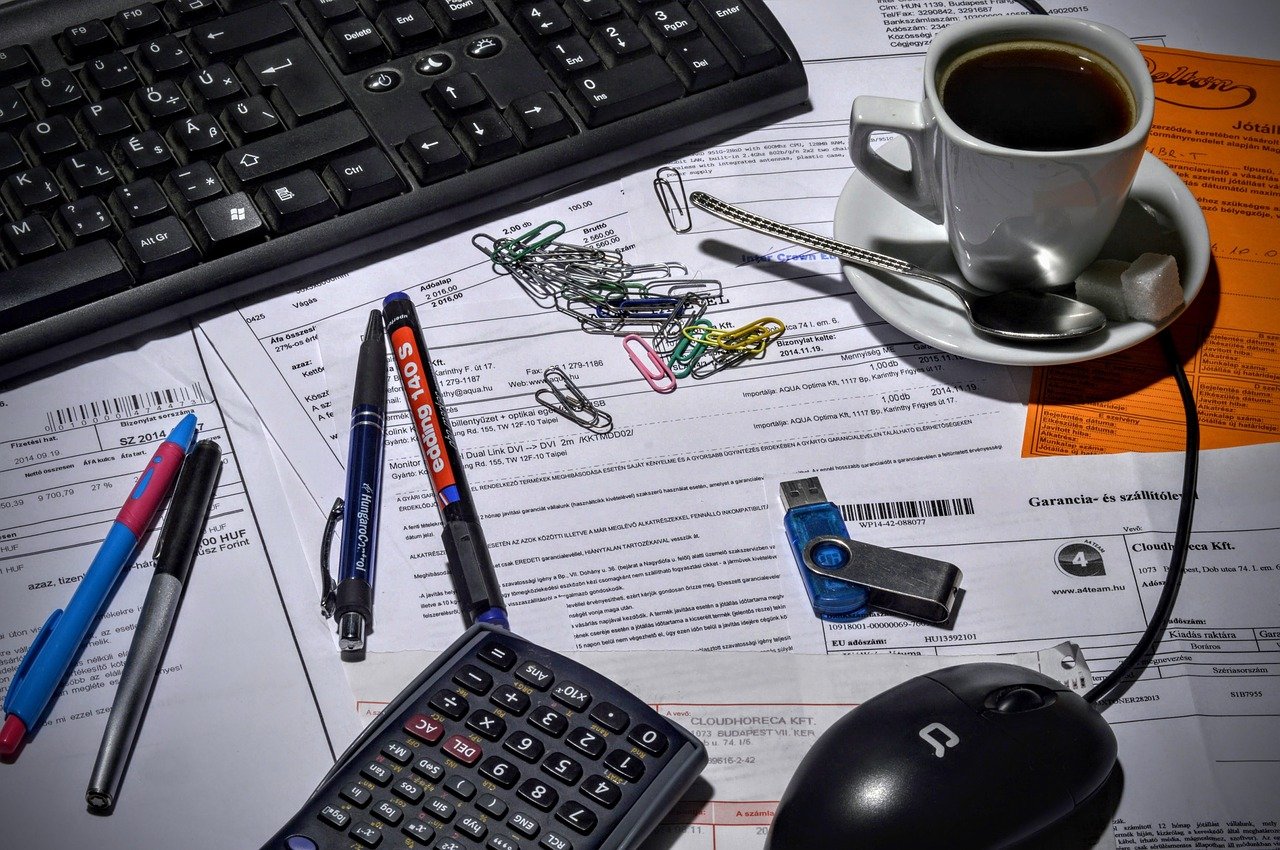4 Benefits to Hiring a Bookkeeper for Your Business - office stationery, calculator, computer, and a cup of coffee on a cluttered desk