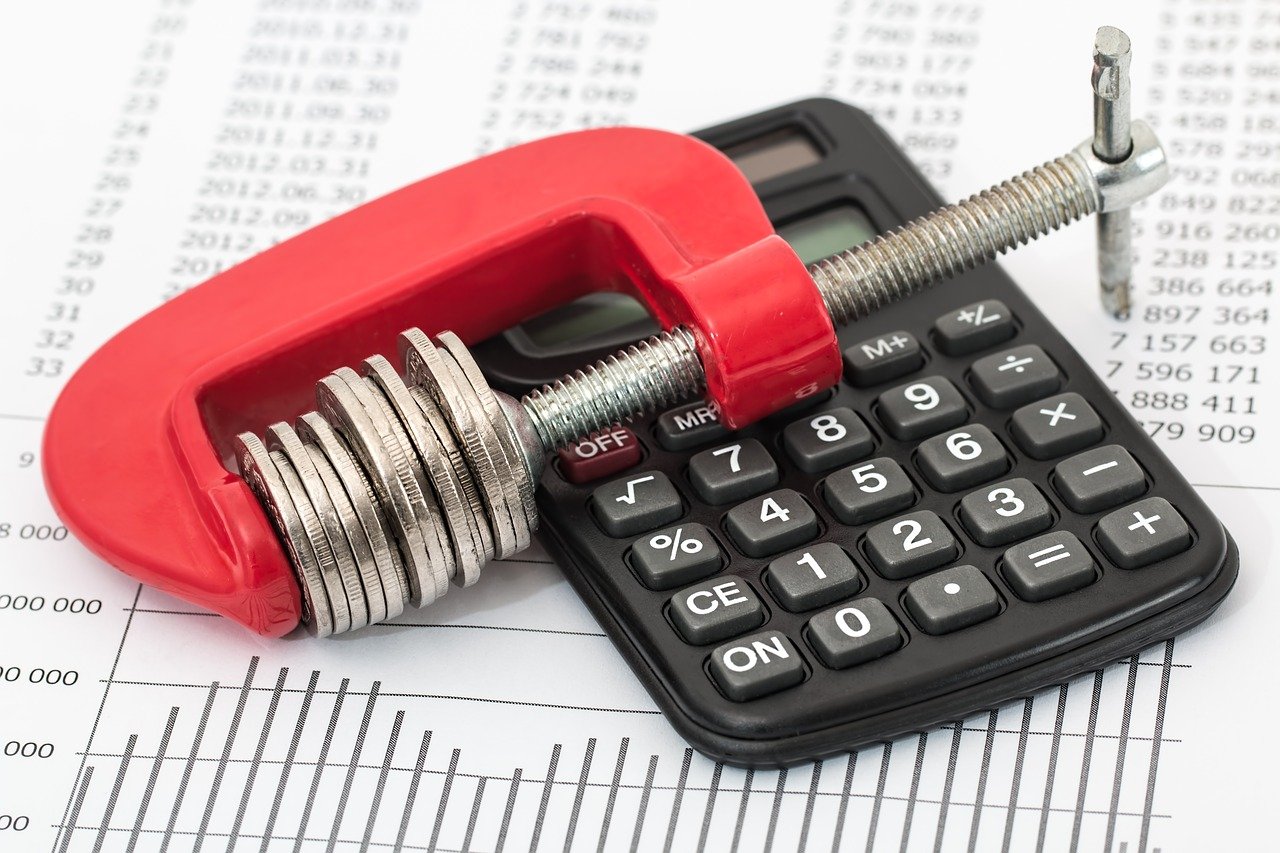 4 Money Saving Tips for Business Owners - A stack of coins held in a clamp on top of a calculator and a business report.